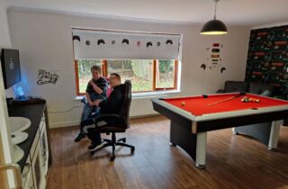 Games room with pool table in the centre, two gaming chairs with tv and console in the corner. The room is decorated with game-themed wallpaper and blinds with gaming controller illustrations.