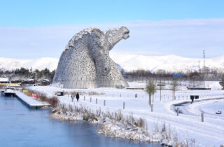 Scenic shot of the Kelpies at Falkirk's Helix, covered in snow. Ochil hills, also dusted in snow, in the background, and the canal in the foreground.